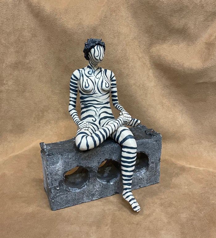 Stoneware ceramic figure of a woman painted with zebra print markings along her face and body sitting with one leg across her thigh as her other leg dangles over a brick that she is sitting on.